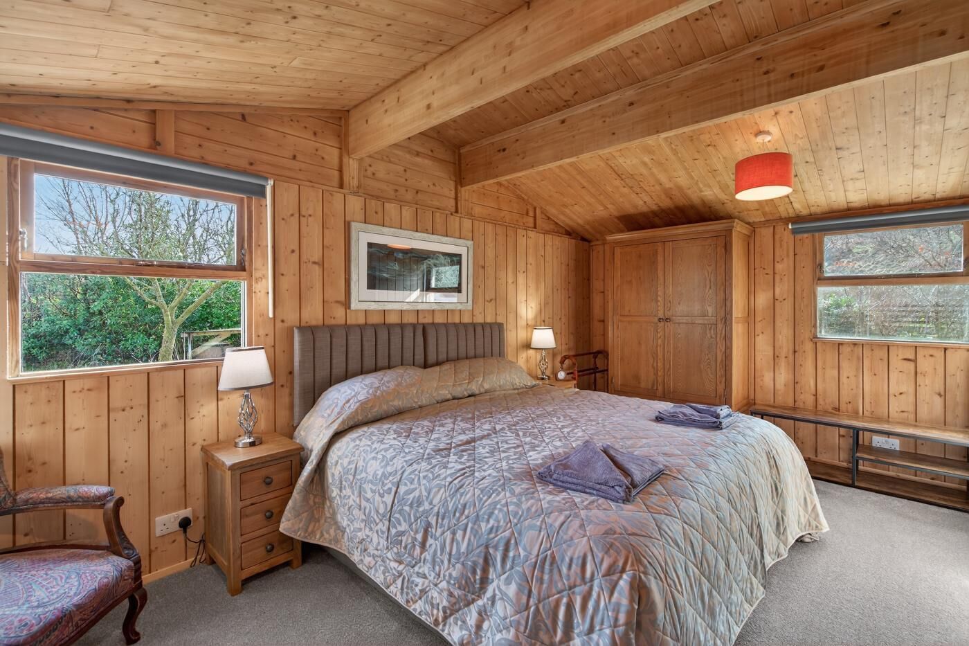 A wooden-paneled bedroom with a bed, bedside tables, a chair, and a window.