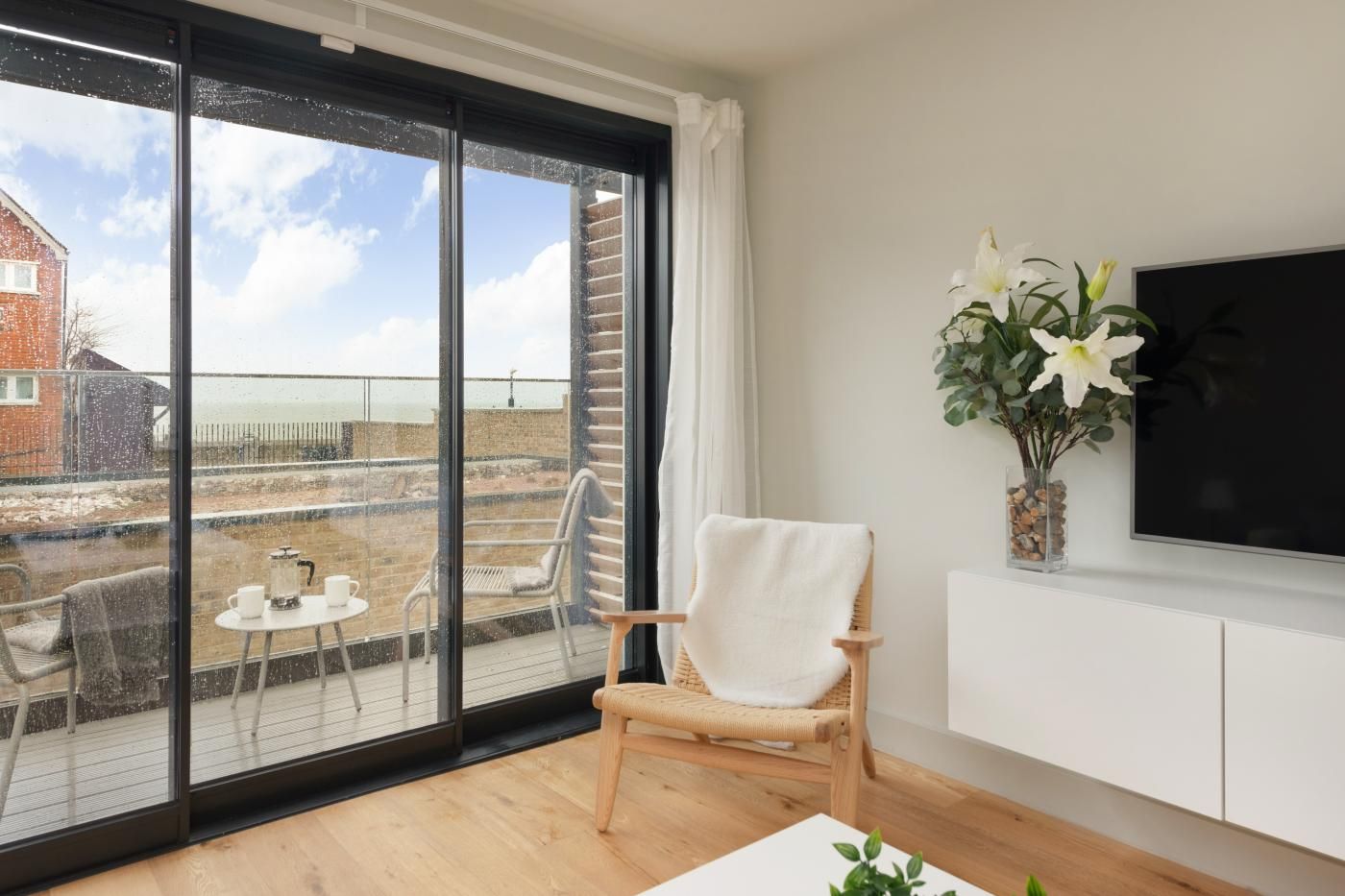A modern living room with a large sliding glass door leading to a balcony, a white armchair, a small table with a coffee pot, and a flat-screen TV mounted on the wall.