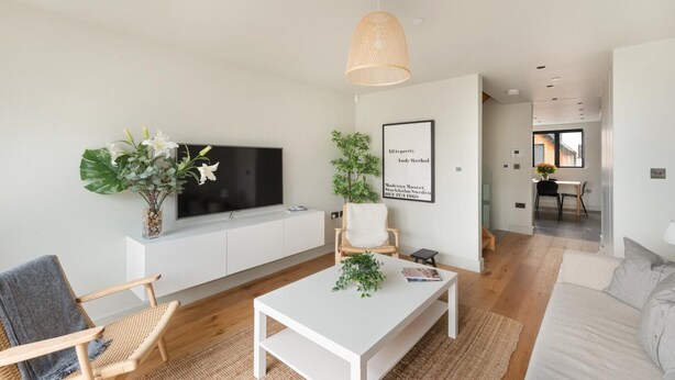 Ett modernt vardagsrum med en platt-TV, ett vitt soffbord, en flätad fåtölj, en vit soffa och en hängande lampa.