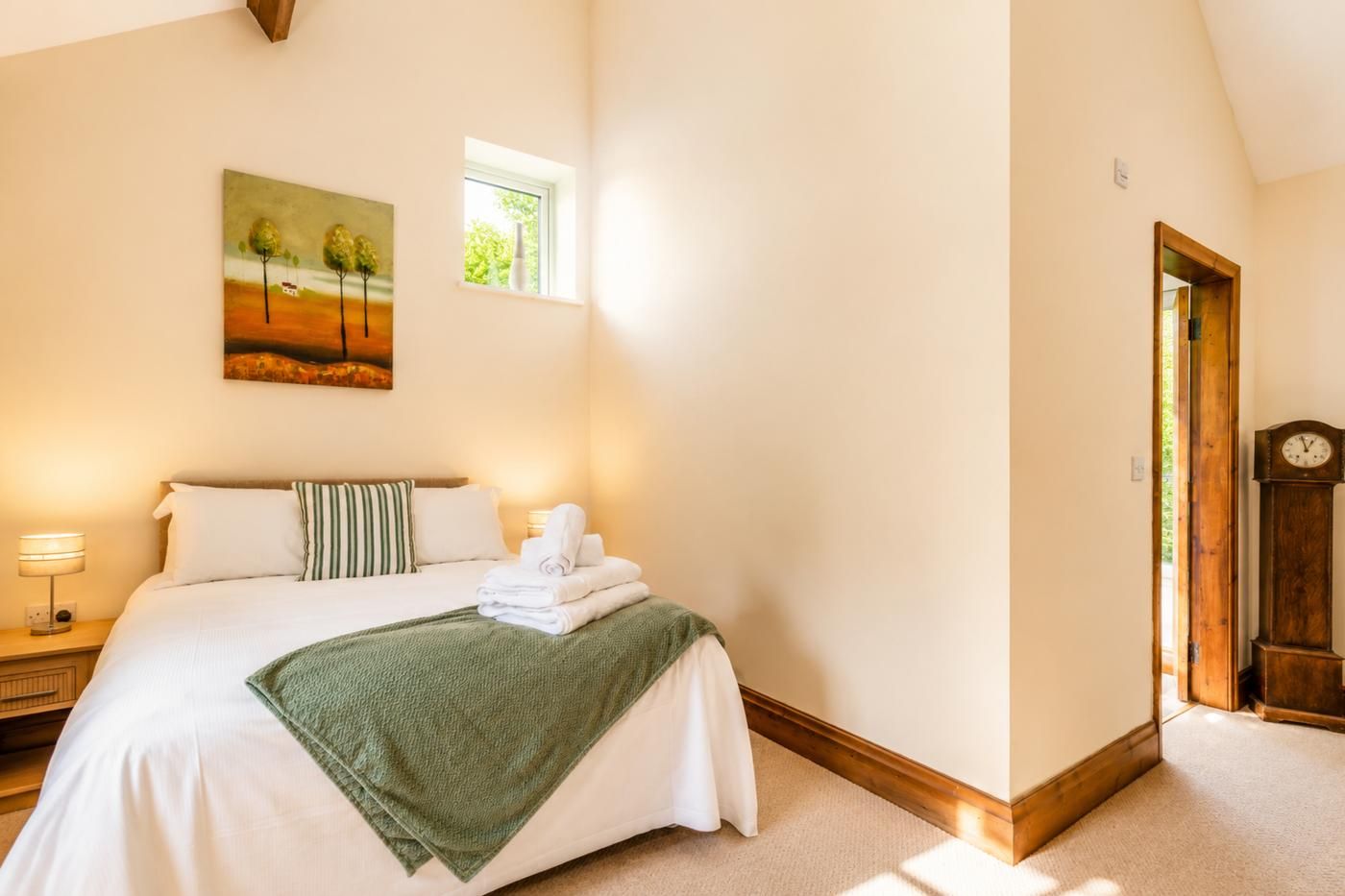 A bedroom with a bed, pillows, a green throw blanket, a bedside lamp, a painting, a window, and a wooden door.