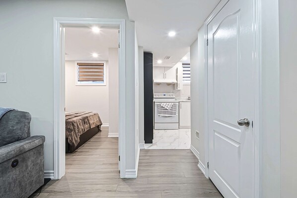 Un pasillo estrecho que da a un dormitorio con cama y sofá gris, una zona de cocina con armarios y electrodomésticos blancos, y una puerta blanca a la derecha.