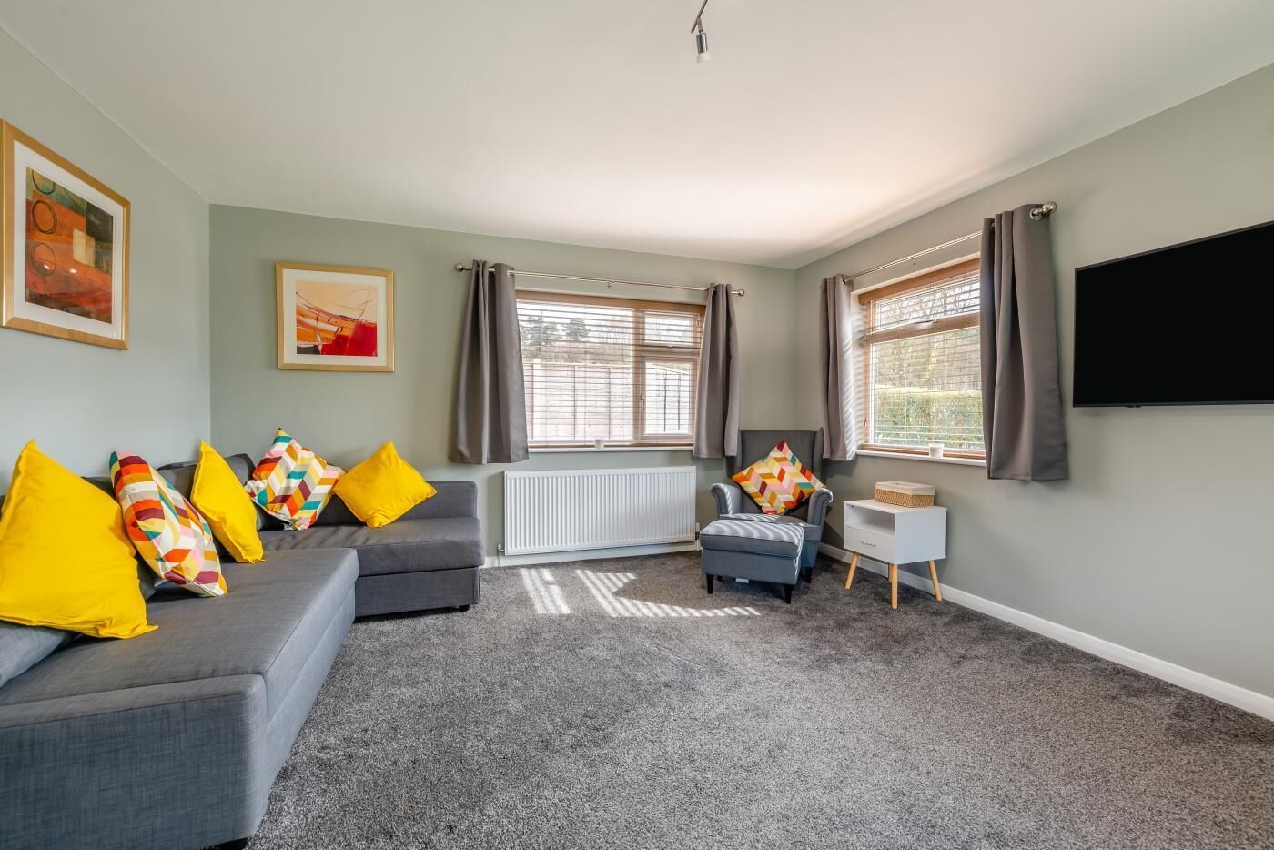 A living room with a grey sofa, yellow pillows, a white side table, and a flat-screen TV.