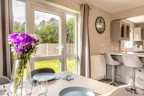 A modern kitchen with a large window, a dining area with a table set for two, and a clock on the wall. - 3 Bed in Matlock Moor (oc-m36349) (Matlock)
