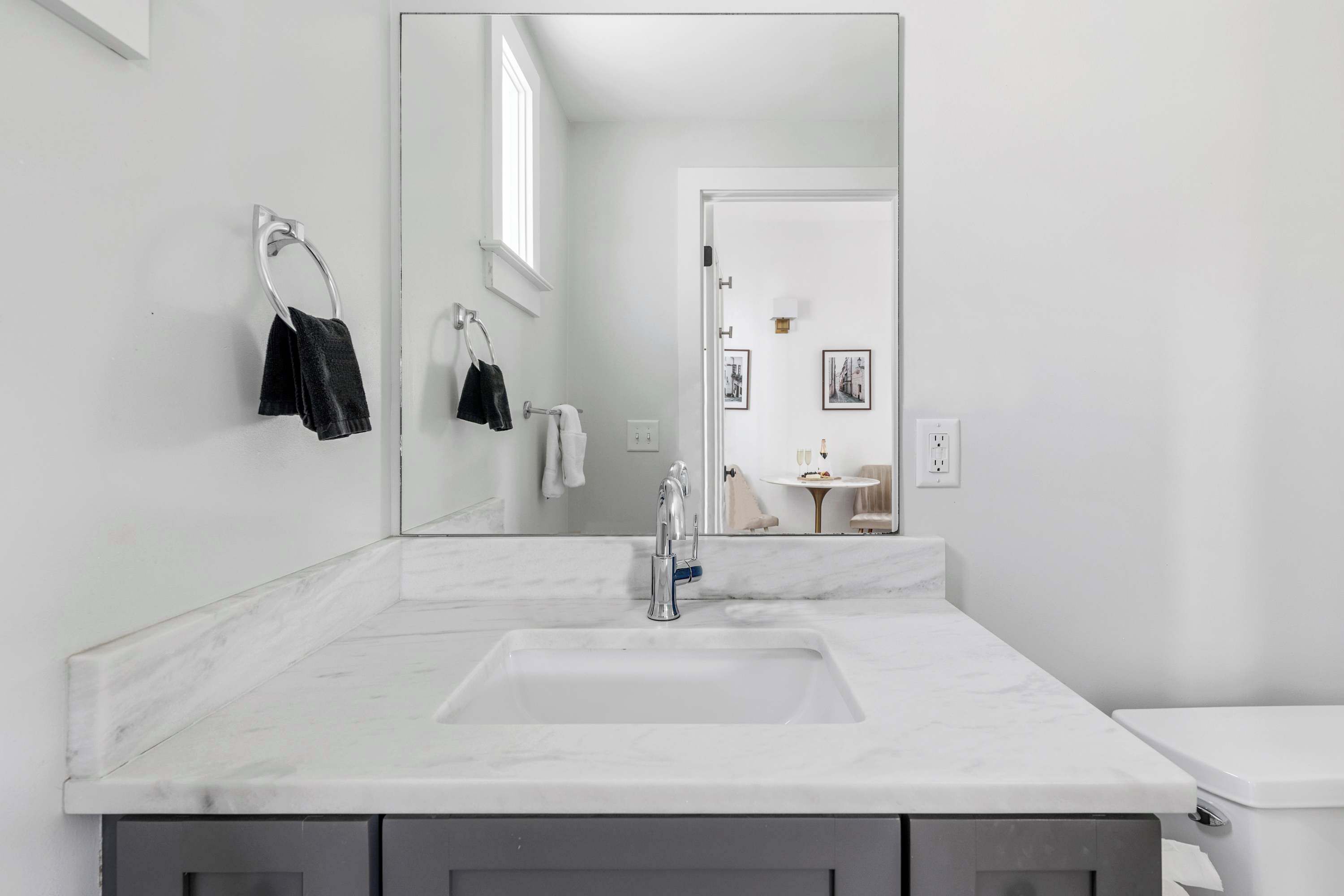 A modern bathroom with a large mirror, a white sink, and a toilet. There are towels hanging on a rack and a small table with a lamp in the background.