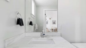 Et moderne badeværelse med et stort spejl, en hvid vask og et toilet. Der er håndklæder hængende på en holder, og et lille bord med en lampe i baggrunden.