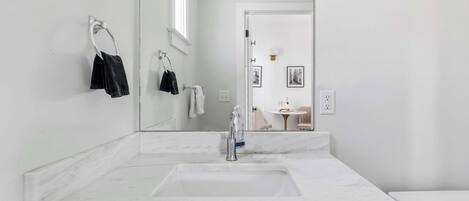 A modern bathroom with a large mirror, a white sink, and a toilet. There are towels hanging on a rack and a small table with a lamp in the background.