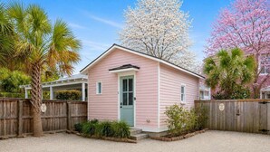 Et pink hus i et etage med en grøn dør, omgivet af et træhegn, grusvej og forskellige planter.