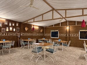 Habitación con techo alto, ventiladores de techo blancos, paredes de madera y alfombra estampada. Se disponen mesas y sillas para comer.