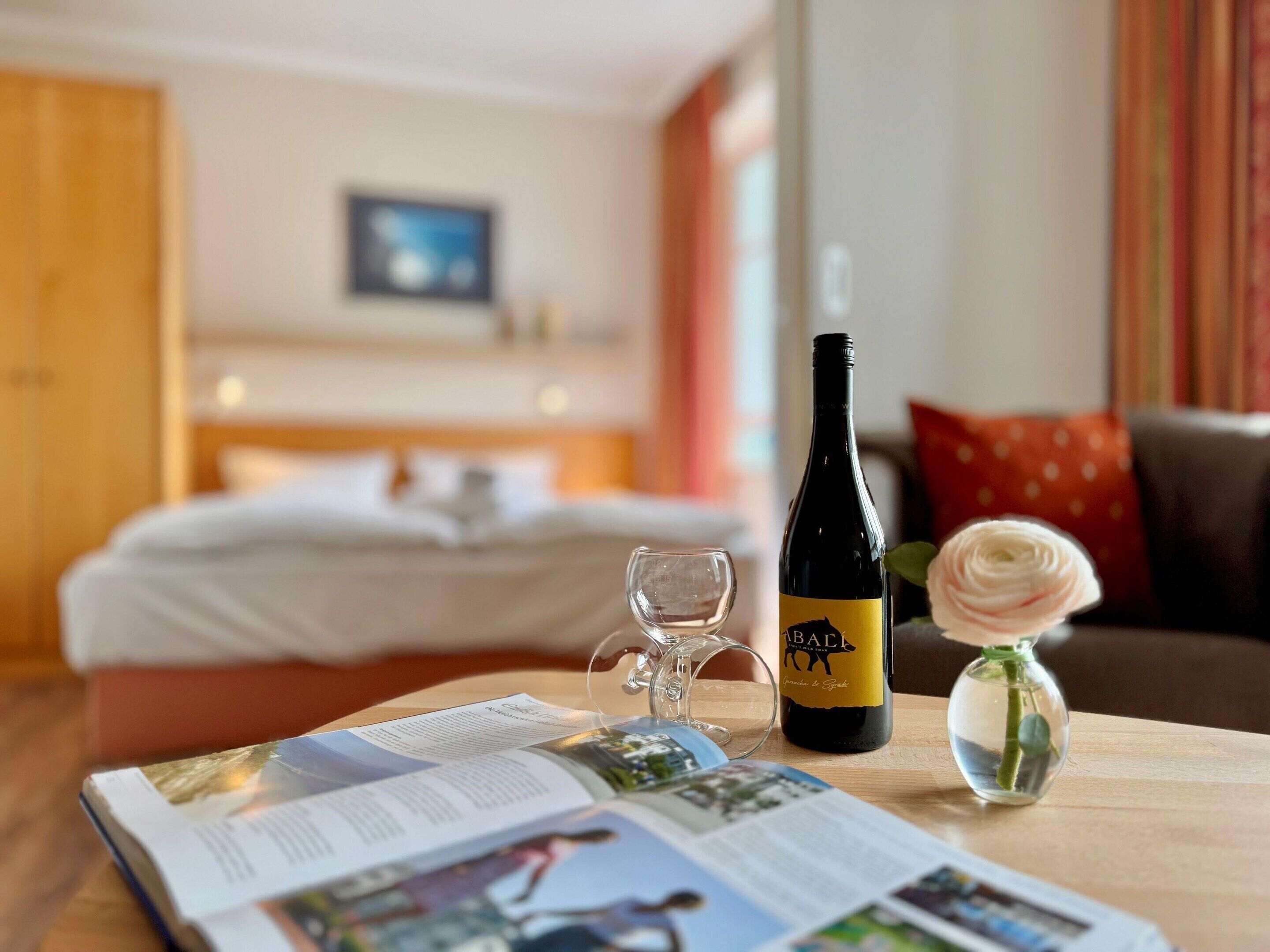 Et hotelværelse med en seng, en vinflaske, et glas og et magasin på et træbord.