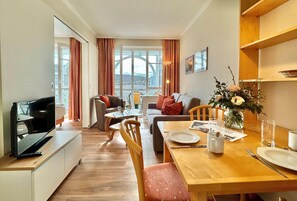 A modern living room with a flat-screen TV, a sofa, a dining table set for two, and a large window with curtains. - Ferienwohnung im Seepark Sellin mit Balkon (Sellin)