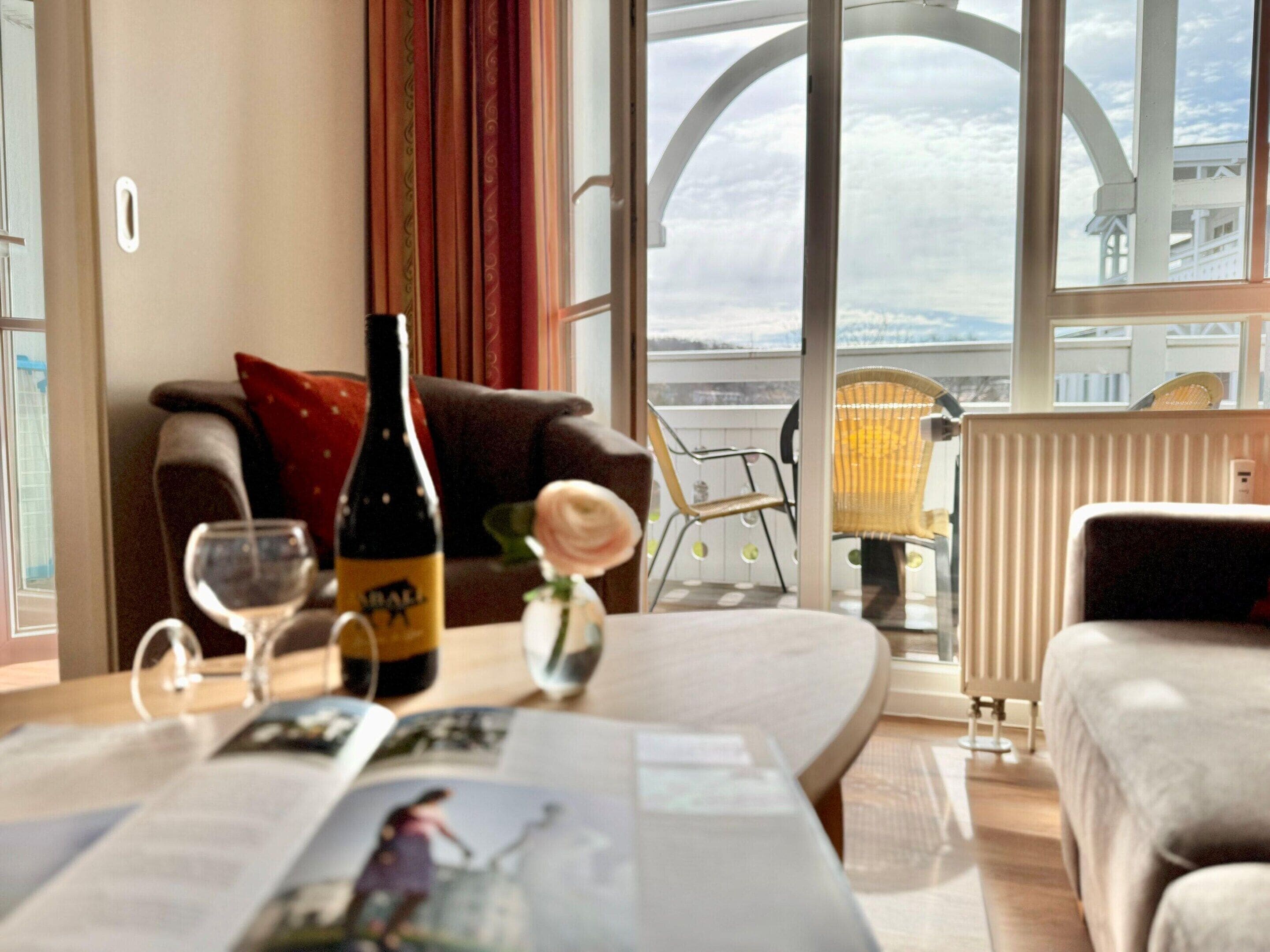 A room with a large window, a table with a bottle and glasses, a sofa, and a chair.