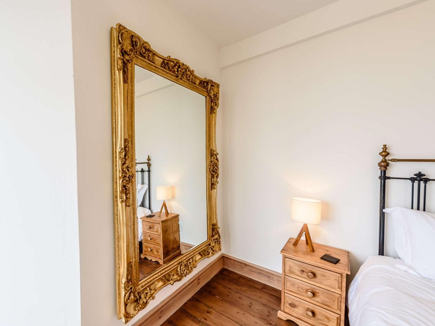 A bedroom with a large ornate mirror, a wooden nightstand with a lamp, a bed with a black metal frame, and a wooden dresser.