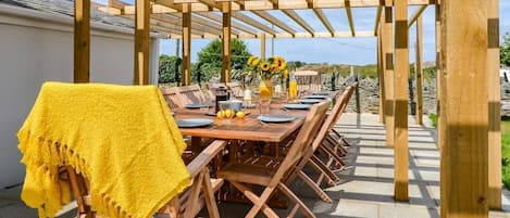 Una pérgola de madera al aire libre con una mesa de comedor preparada para seis personas, rodeada de sillas.