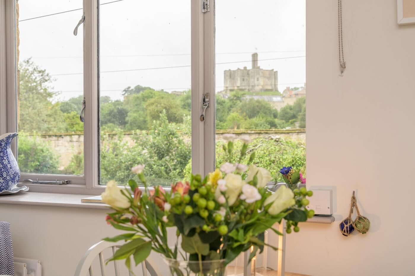 A room with a large window, a vase with flowers, a blue patterned pitcher, and a wall-mounted light fixture.