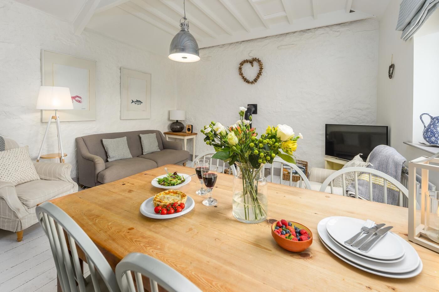 A dining area with a wooden table, chairs, a vase of flowers, plates of food, and a television in the background.