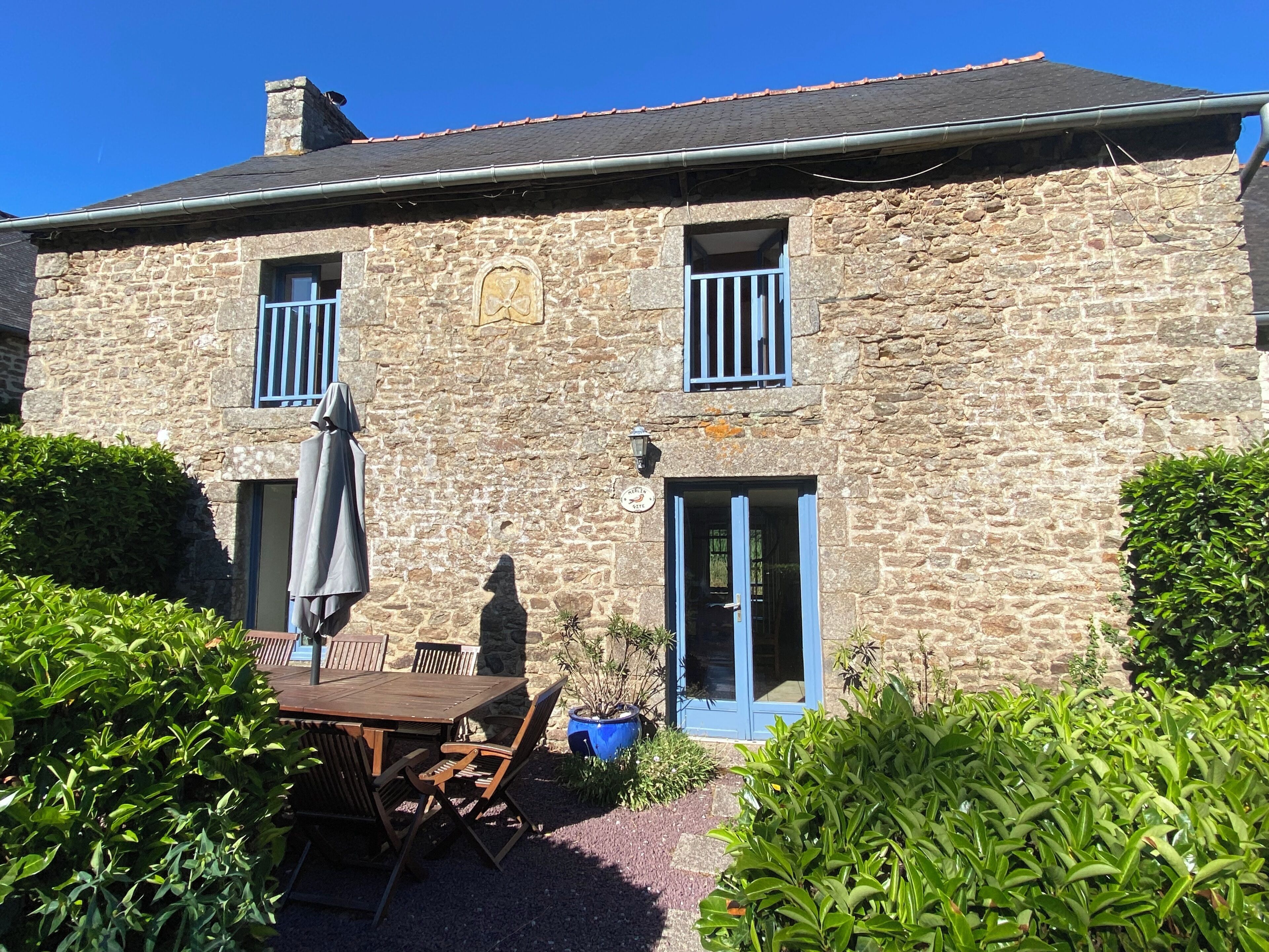 Een stenen gebouw met een houten tafel en stoelen buiten, een blauwe deur en een raam met een traliespatroon.