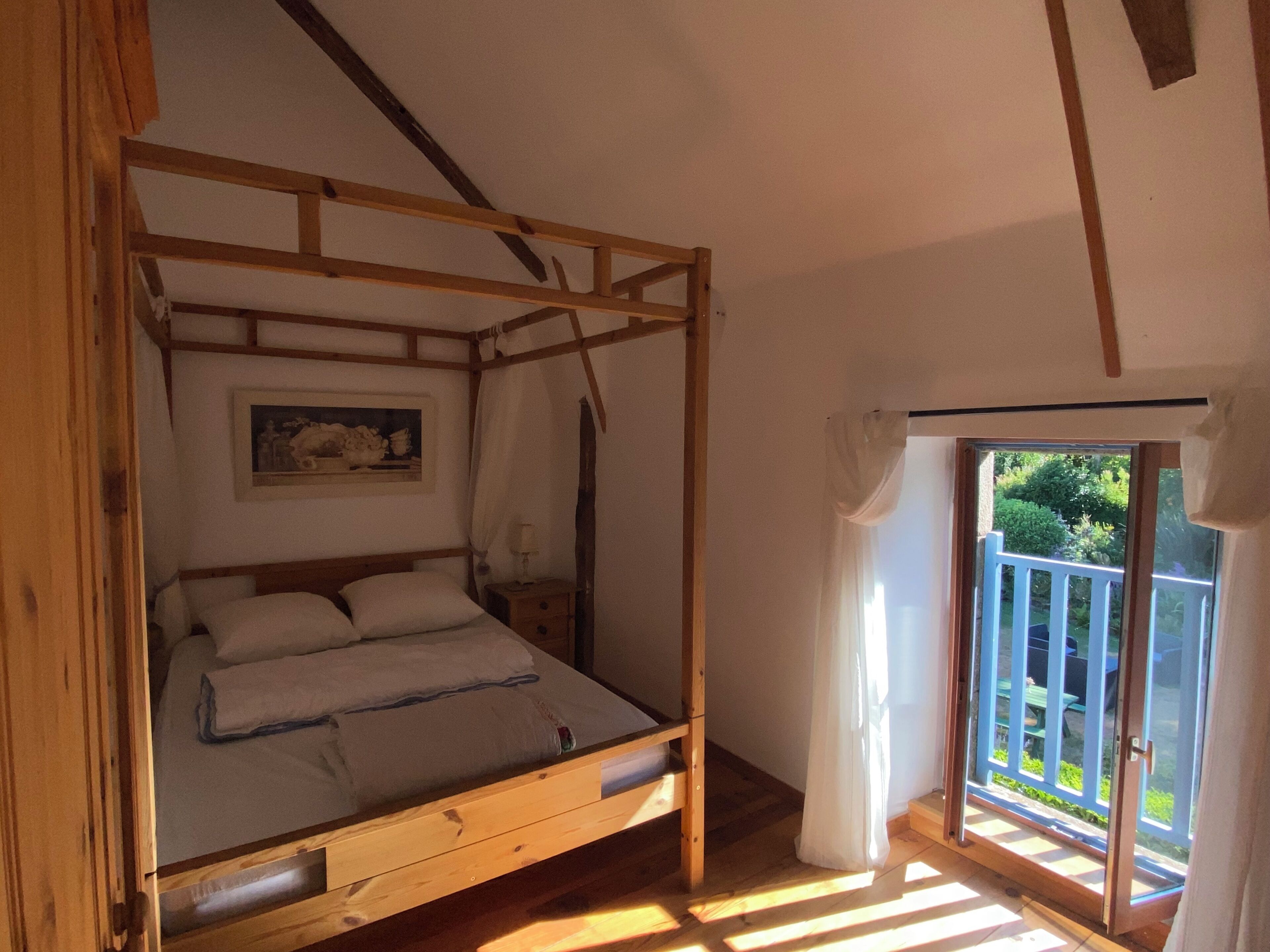 Een houten hemelbed met nachtkastje, een lijst met foto aan de muur en een balkon met uitzicht op groen.