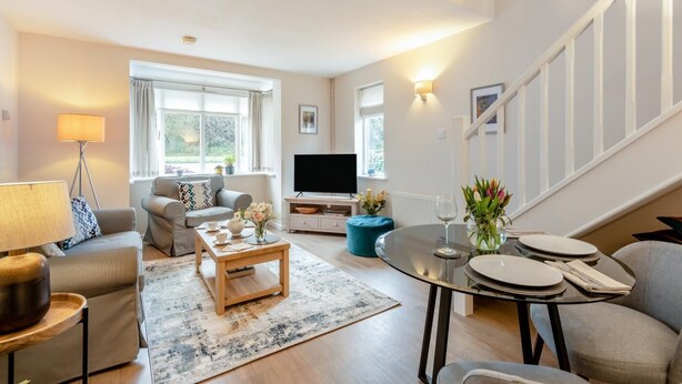 A modern living room with a sofa, coffee table, and TV, a dining area with a table set for two, and a staircase.
