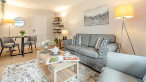 A modern living room with a grey sofa, a coffee table with a book, a dining table with chairs, and a floor lamp.