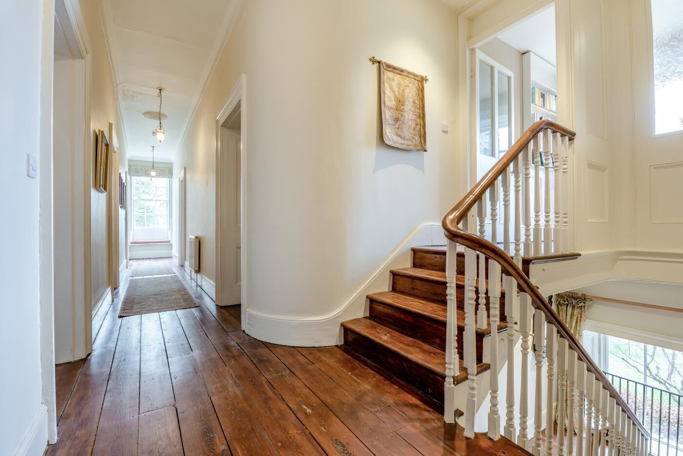 A hallway with wooden flooring, a staircase with white railings, and a hanging tapestry.