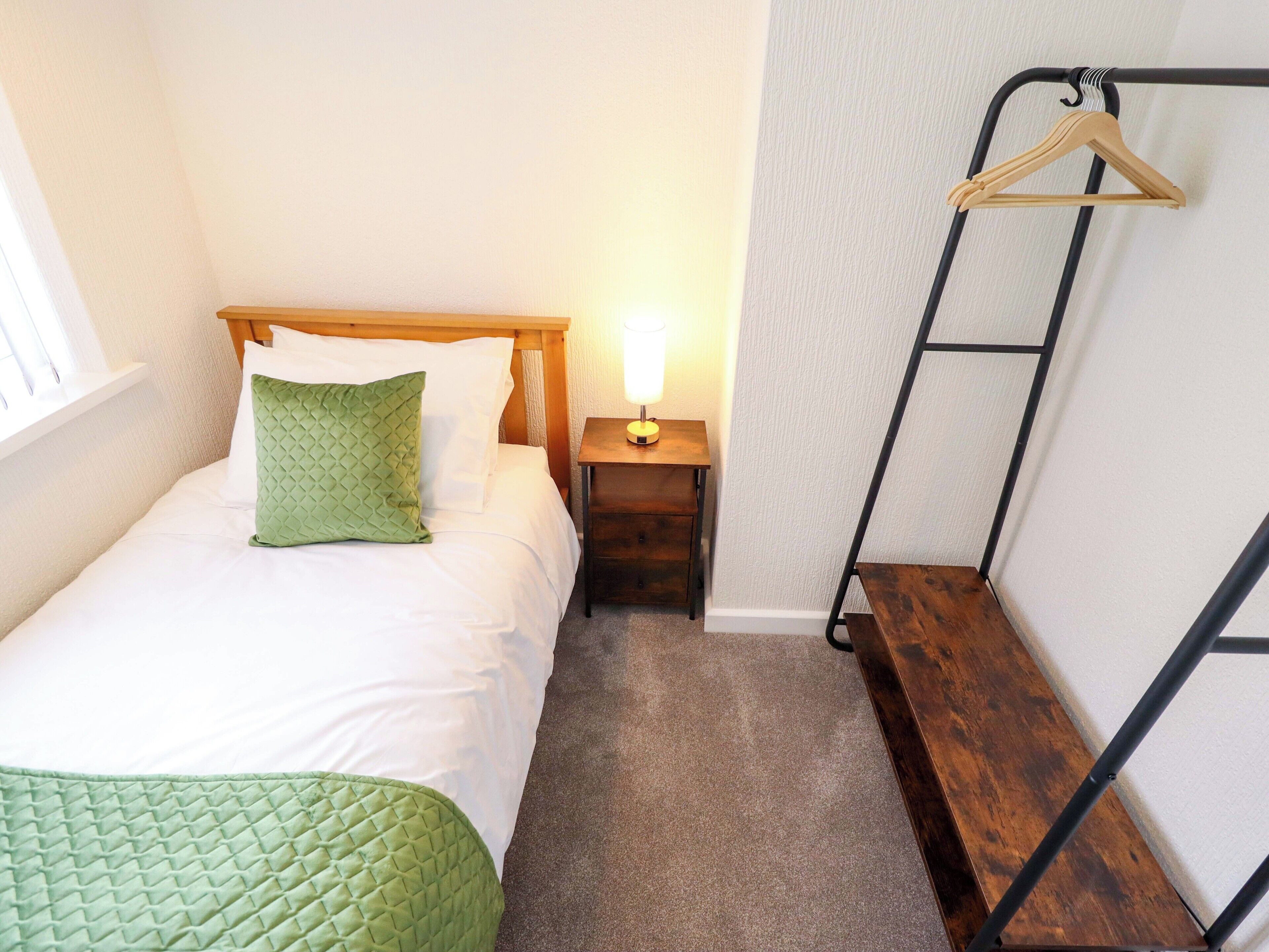 A single bed with a wooden headboard, a green pillow, a bedside table with a lamp, and a wooden bench.