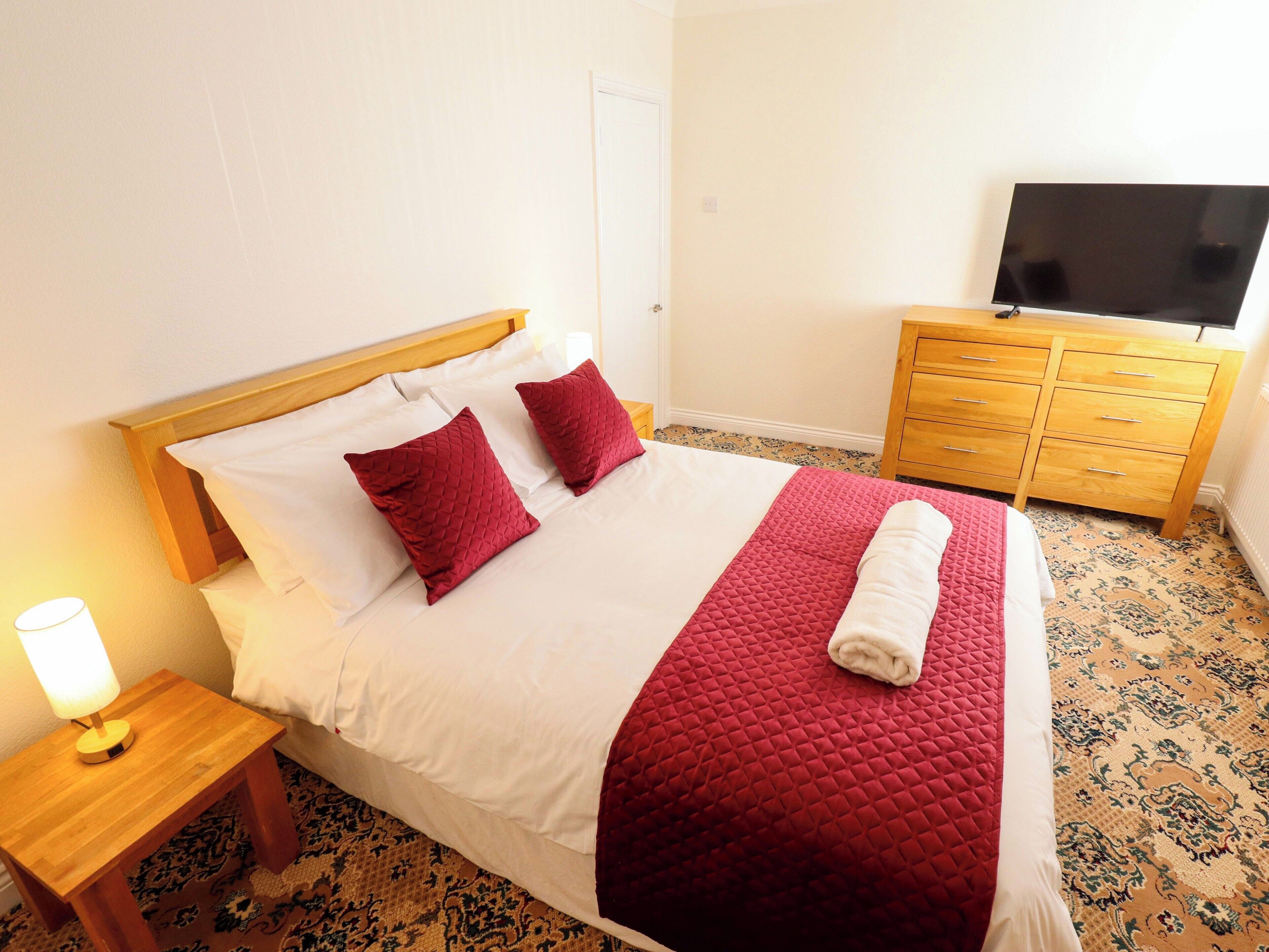 A neatly made bed with a red quilt, a wooden nightstand with a lamp, a wooden dresser with a flat-screen TV, and patterned carpet.