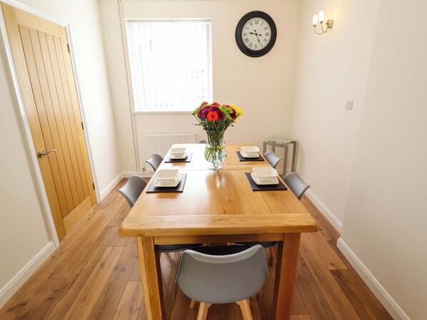 Un comedor con una mesa de madera, sillas, un florero con flores, un reloj en la pared y una ventana con persianas.