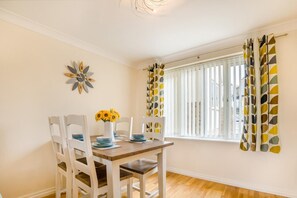 A dining area with a table set for four, white chairs, a vase with sunflowers, and a window with vertical blinds. - 2 Bed in Broadstairs (oc-b39035) (Broadstairs)