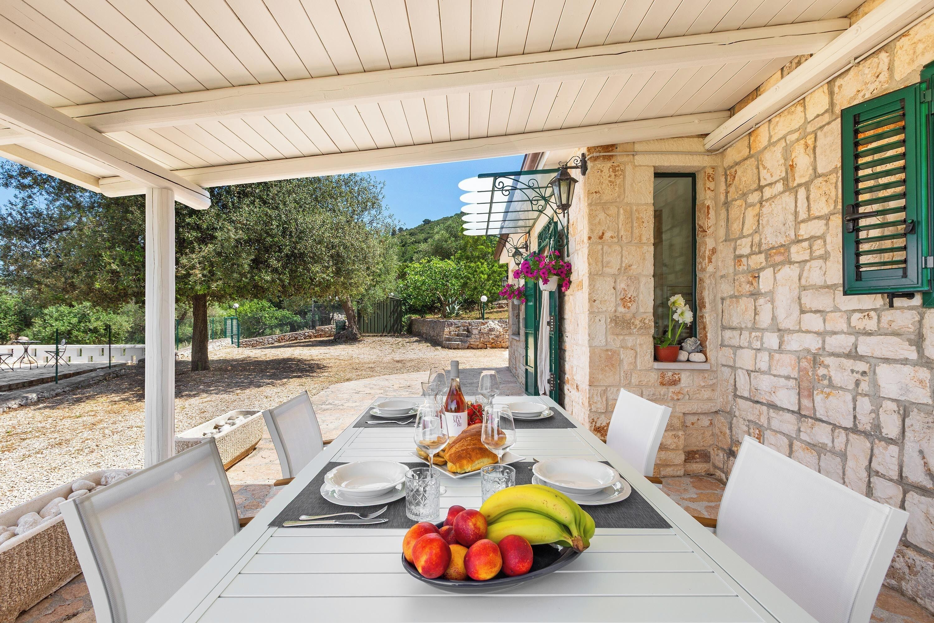 A covered outdoor dining area with stone walls, a table set with plates, glasses, and a fruit bowl.