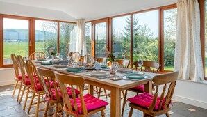 Een eetkamer met een grote tafel gedekt voor zes personen, houten stoelen met roze kussens, en grote ramen met uitzicht op groen.