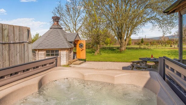 Een hottub met een houten terras, een klein houten gebouwtje met schoorsteen en een gazon met bomen.