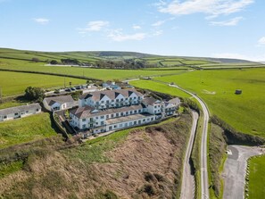 A large, multi-story building with a symmetrical facade, surrounded by green fields and a winding road. - 2 Bed in Putsborough (oc-s40237) (Croyde)