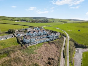 A large, multi-story building with a symmetrical facade, surrounded by green fields and a winding road. - 2 Bed in Putsborough (oc-s40237) (Croyde)