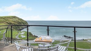 Ein Balkon mit Tisch für zwei Personen, mit Blick auf eine Küstenlandschaft, Meer und einen grasbewachsenen Hügel.