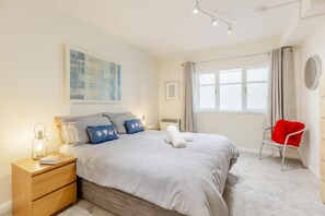 A bedroom with a bed, bedside tables, a chair, a window with curtains, and artwork on the wall. - 2 Bed in Putsborough (oc-s40237) (Croyde)