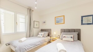 Ein Doppelzimmer mit zwei Betten, jeweils mit Kissen und einem gerollten Handtuch. Nachttische mit Lampen, ein Fenster mit Jalousien und gerahmte Bilder an den Wänden.