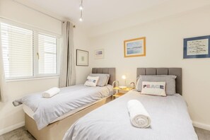 A double room with two beds, each with a pillow and a rolled towel. There are bedside tables with lamps, a window with blinds, and framed pictures on the walls. - 2 Bed in Putsborough (oc-s40237) (Croyde)