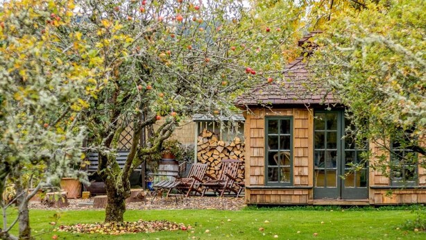 Una cabaña de madera rústica con un pequeño porche, una pila de leña y un árbol lleno de manzanas.