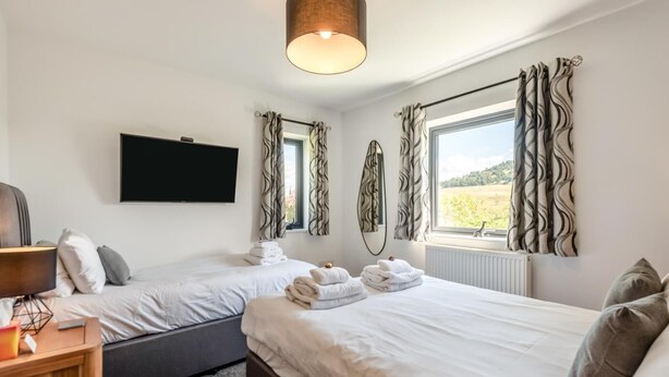 A hotel room with two beds, a flat-screen TV, a lamp, and a view of the outdoors. - 3 Bed in Penrith (oc-89427) (Hutton)