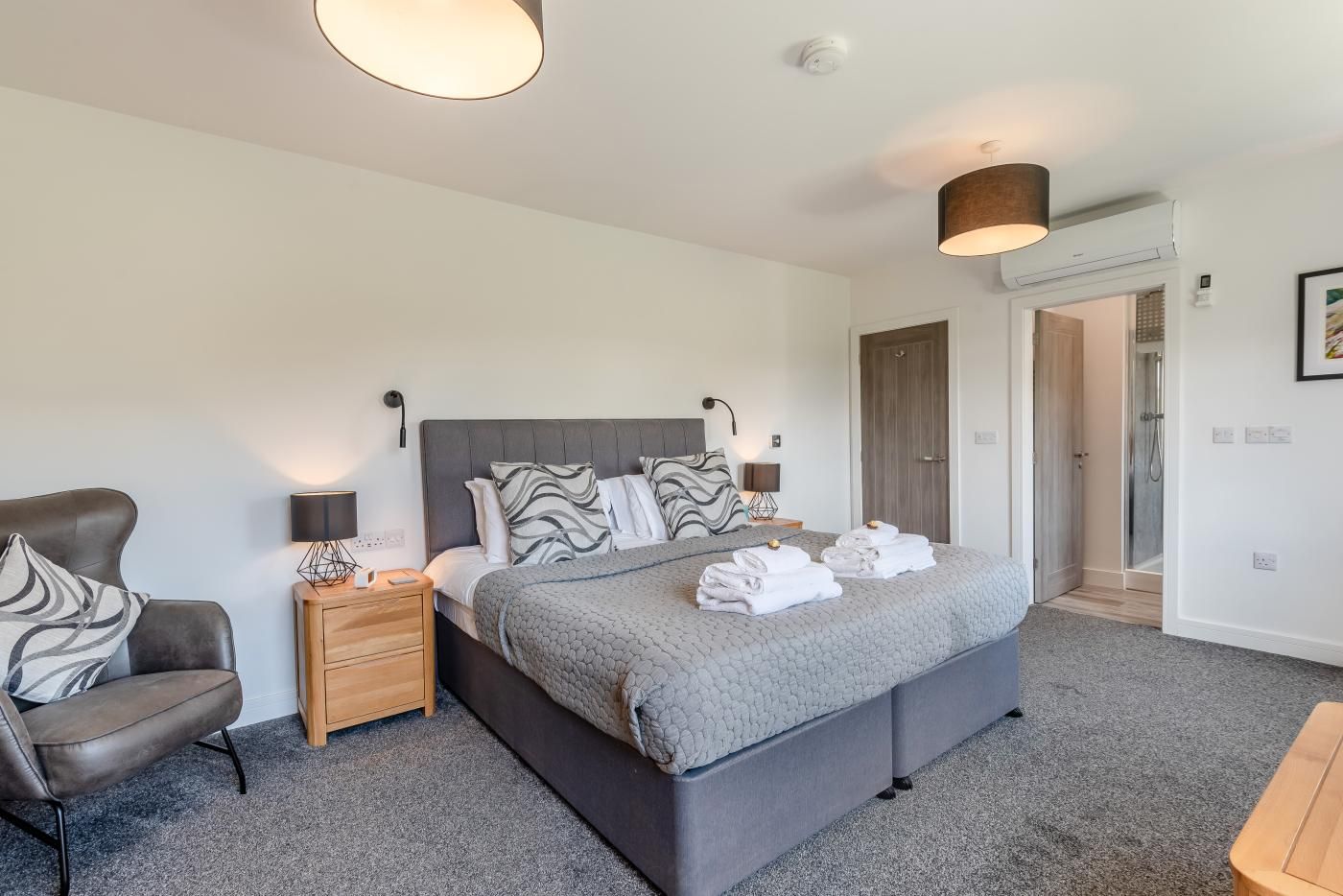 A modern bedroom with a large bed, bedside tables, a chair, and a shower area.
