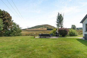 Une cour arrière verdoyante avec un banc, un barbecue et une maison en arrière-plan.