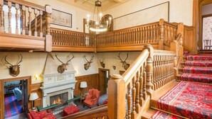 A grand interior with a fireplace, plush seating, a staircase with ornate railings, and mounted animal heads on the walls. - 12 Bed in Tomatin (oc-ca203) (Woodend)