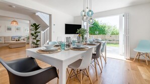 Un coin repas avec une table blanche dressée pour six personnes, entourée de chaises. Un téléviseur est fixé au mur, et un escalier mène à une autre pièce.