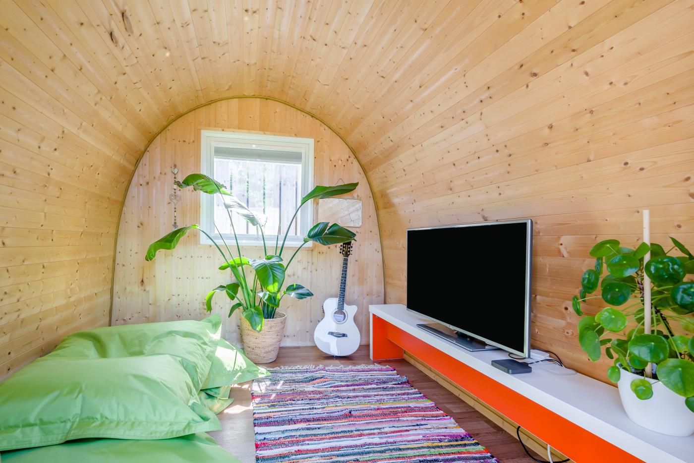 A cozy room with a curved wooden ceiling, a bed with green pillows, a television on a white stand, and a guitar leaning against the wall.