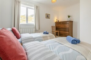 Ein Schlafzimmer mit zwei Betten, einem Holzkleiderschrank, einem Sessel, einem Fenster mit Vorhängen und einer kleinen Pflanze.
