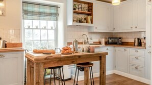 Une cuisine avec un îlot central en bois, des meubles blancs, une fenêtre avec des stores et un luminaire suspendu.