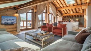 Een ruime woonkamer met een hoge plafond, houten balken, een televisie, een bank, een salontafel en uitzicht op bergen door grote ramen.