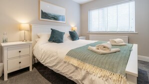 Ein ordentlich eingerichtetes Schlafzimmer mit Bett, Nachttischen, einem Fenster mit Jalousien, einem Heizkörper und einem gerahmten Bild an der Wand.