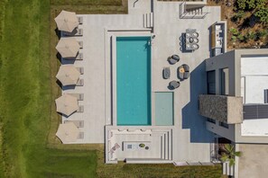 Vista aèria d'un edifici modern amb una piscina rectangular, diverses gandules i ombrelles.