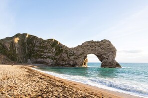 A beach with a natural rock arch formation, clear skies, and calm sea. - 3 Bed in Piddlehinton (oc-c40254) (Piddlehinton)